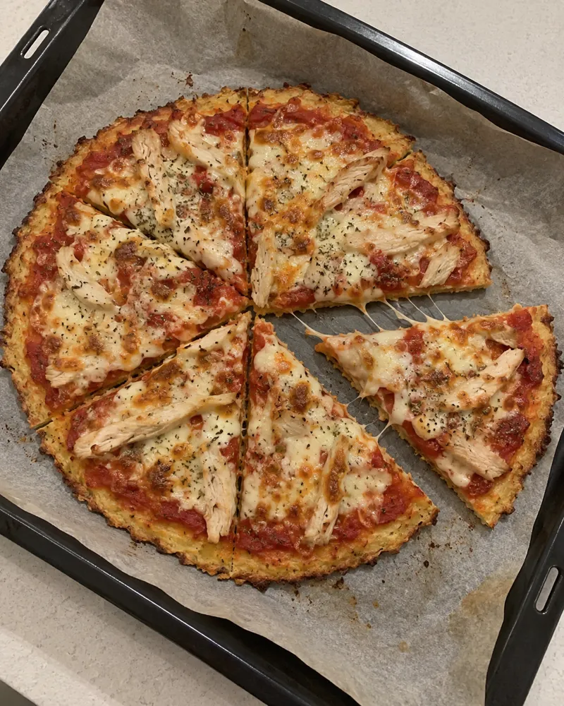 Pizza fina de base de arroz dorada en una bandeja, con tomate, mozzarella fundida, tiras de pollo y orégano, cortada en 6 trozos