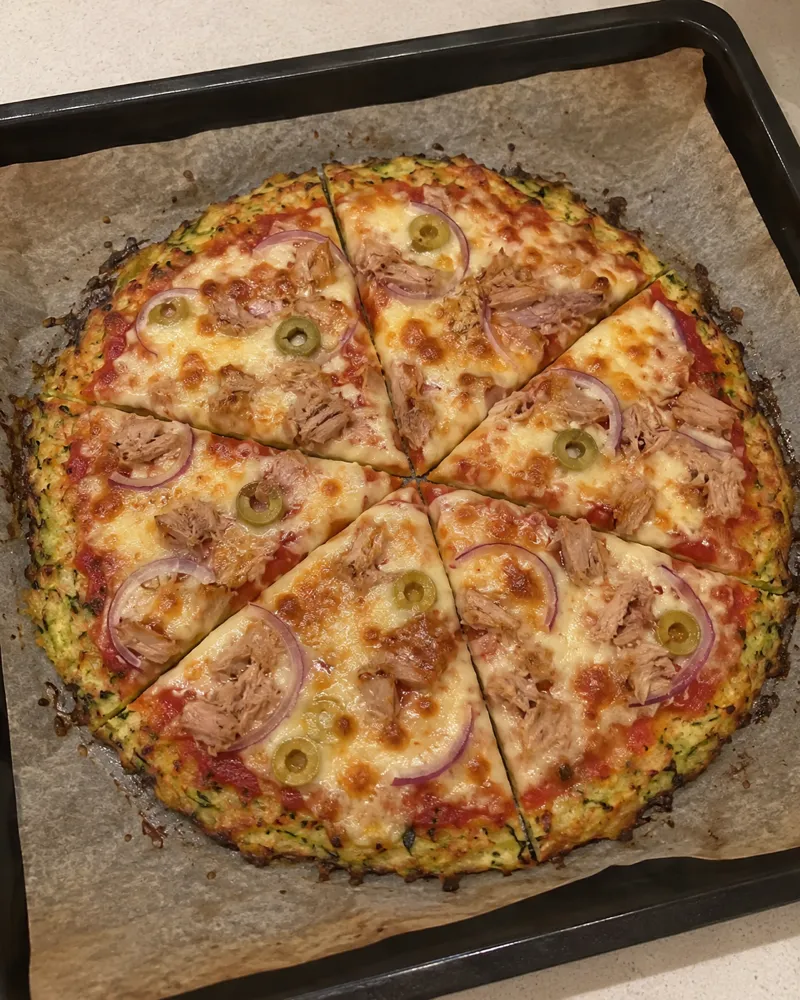Pizza de base de calabacín dorada con tomate, atún, aros de cebolla roja, aceitunas verdes y mozzarella fundida, cortada en 6 trozos